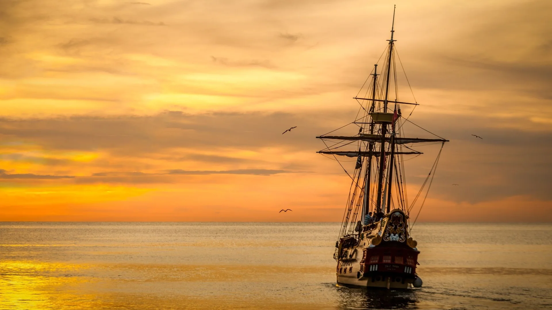 A replica galleon under sail on a calm sea, its masts black against an orange sunset sky