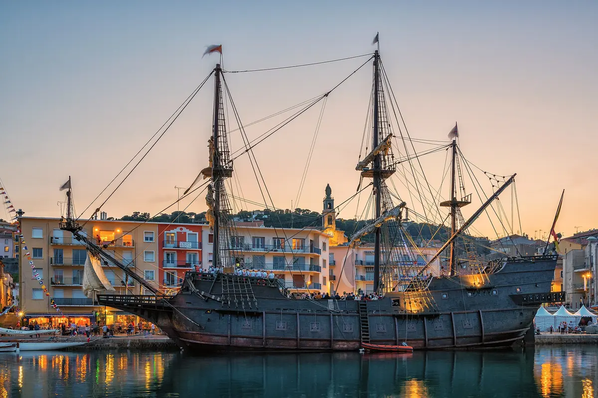 A replica galleon with tall masts moored in a harbour at dusk, with lit waterfront buildings behind it