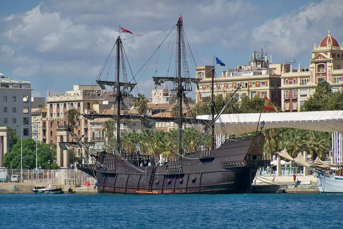 A dark wooden replica galleon with tall masts moored in front of a row of grand harbour buildings