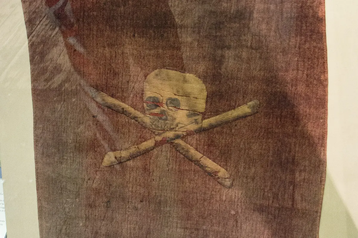A faded red cloth flag with a pale skull above crossed bones, displayed behind glass