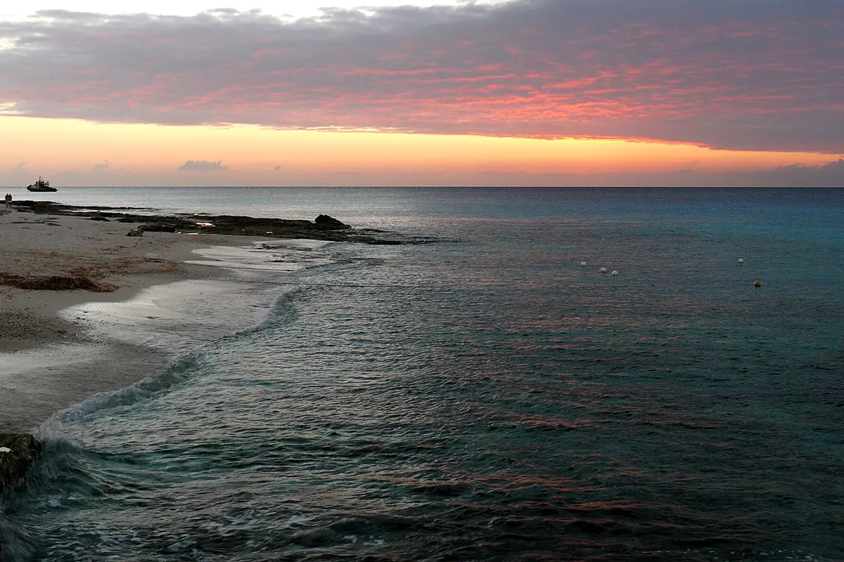 A pink and orange sunset sky over calm water and a rocky shoreline on Cozumel's western shore
